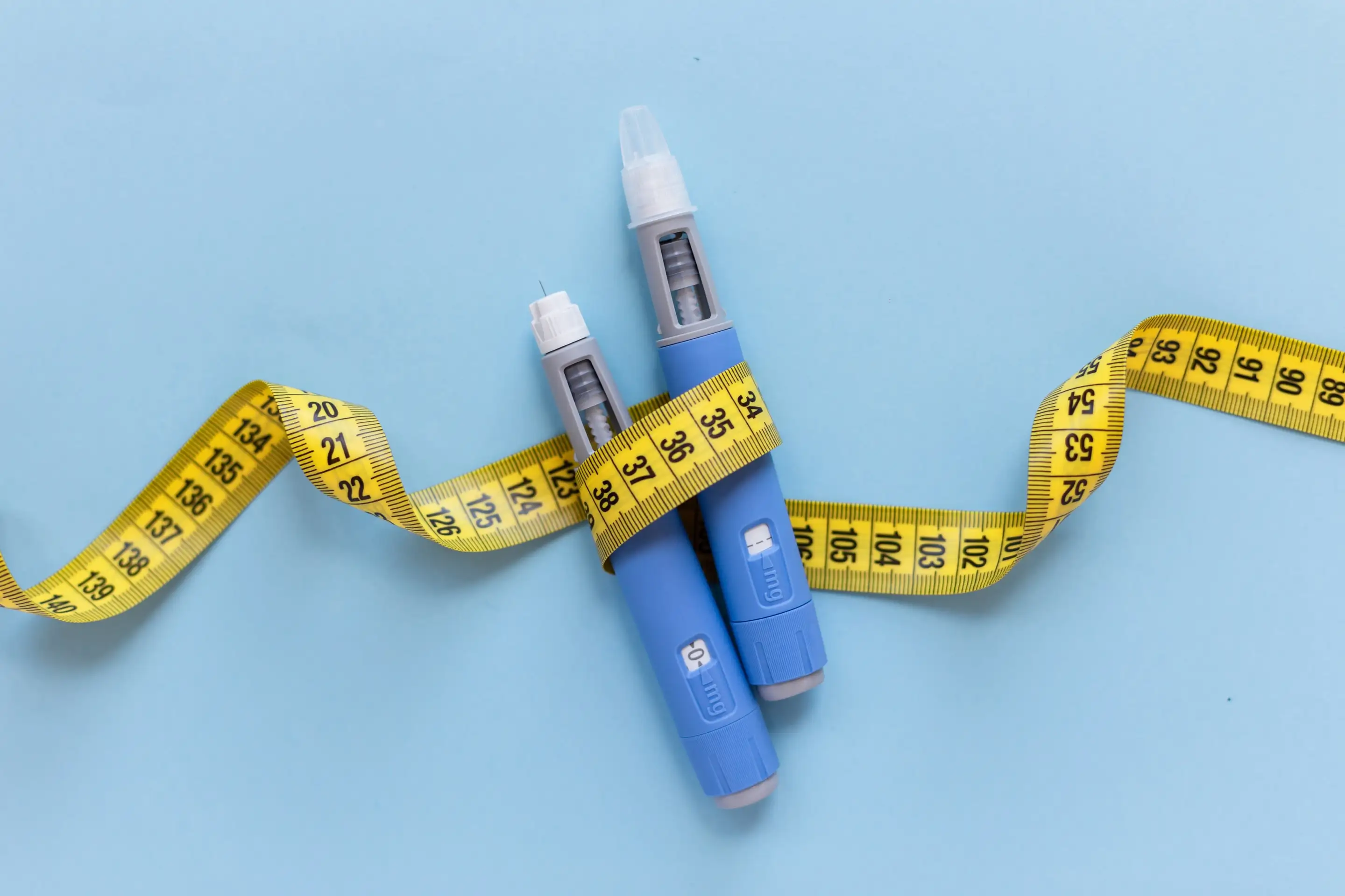 Two injection pens beside a tape measure on a blue surface